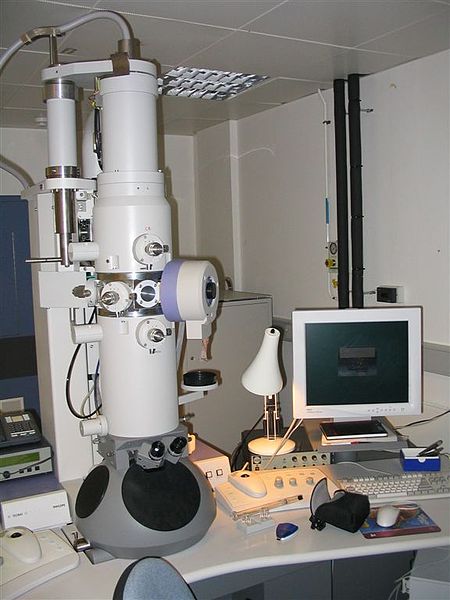 A photograph of an electron microscope. It is a large white tube positioned vertically on a large desk. Next to the microscope on the desk is a computer, which is connected to the microscope.